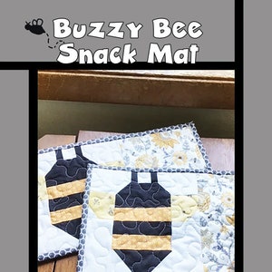 May include: Two quilted bee-themed snack mats with a white background, black and yellow bee design, and gray and white floral fabric border. The mats are on a wooden surface. The text "Buzzy Bee Snack Mat" is at the top of the image. The text "Tricia Lynn Maloney Orphan Quilt Designs for Villa Rosa Designs" is at the bottom of the image.