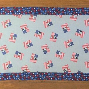 May include: A rectangular table runner with a patriotic theme. The center features a light blue fabric with a repeating pattern of American flags. The edges and ends are trimmed with a blue fabric with red and white floral designs.
