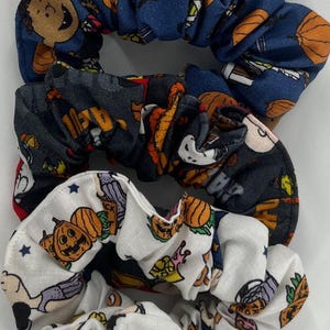 May include: A stack of five fabric hair scrunchies featuring various Halloween-themed prints. The designs include cartoon characters dressed in costumes, orange pumpkins, jack-o'-lanterns, and bats on gray, blue, black, and white backgrounds.