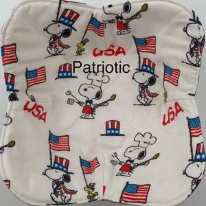 May include: A white, quilted oven mitt featuring a repeating pattern of Snoopy characters in patriotic attire, American flags, and the word "USA" in red. The word "Patriotic" is also printed on the mitt.
