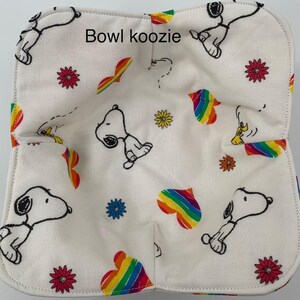 May include: A white bowl koozie with a Snoopy pattern, rainbow hearts, and flowers. The koozie is square-shaped and has a quilted design. The text "Bowl koozie" is printed on the top.