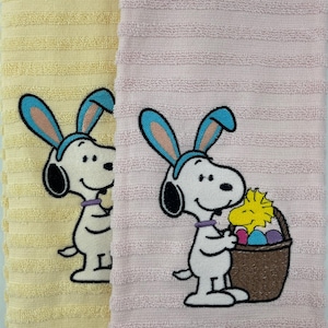 May include: Two Easter-themed hand towels. One is pale yellow, the other is pink. Each features an embroidered Snoopy wearing bunny ears. One Snoopy holds a basket of Easter eggs and a chick. The towels have a ribbed texture.