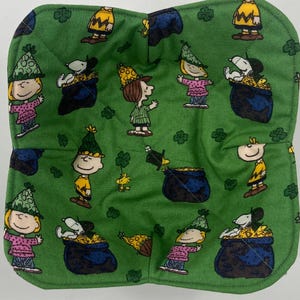 May include: Green fabric bowl cozy featuring cartoon characters, including Snoopy, Charlie Brown, and Lucy, with pots of gold and shamrocks. The cozy is quilted and has a stitched edge. The characters are wearing festive hats, suggesting a St. Patrick's Day theme.