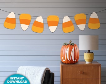May include: A Halloween-themed garland of candy corn shapes hangs above a wooden cabinet. An orange pumpkin with a white "M" sits on the cabinet. The image also includes a lamp and the text "PRINTABLE FALL DECOR".