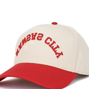 May include: A baseball cap with a cream-colored crown and a red brim. The red text "KANSAS CITY" is embroidered across the front of the cap. The cap has a red button on top.