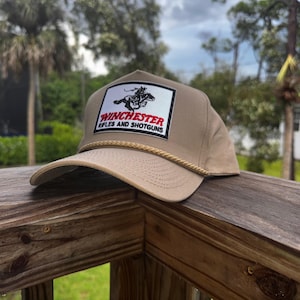 May include: Tan baseball cap with a rope brim. The cap features a rectangular patch with a black and white graphic of a cowboy on a horse, and the words "Winchester Rifles and Shotguns" in red and white.