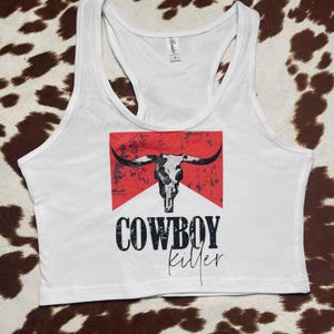 May include: White racerback tank top with a graphic design featuring a bull skull, red background, and the words "COWBOY killer" in black lettering. The tank top has a scoop neck and a racerback design.