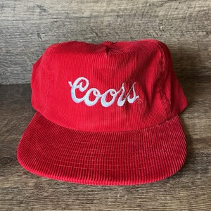 May include: A red corduroy baseball cap with a curved brim. The cap features the word "Coors" embroidered in white script across the front. The hat is a classic style, suitable for casual wear.