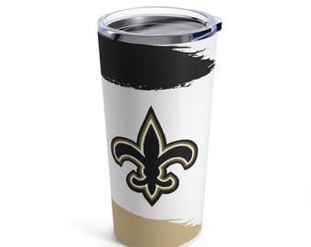 May include: A white stainless steel tumbler with a black and gold fleur-de-lis design. The tumbler has a black and gold brushstroke pattern on the top and bottom.
