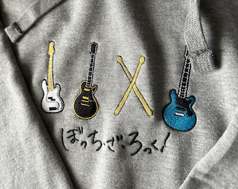 May include: A gray hoodie with embroidered images of a white bass guitar, a black electric guitar, a pair of drumsticks, and a blue electric guitar. The text "(ま?っ)  う  る  !" is embroidered below the guitars.