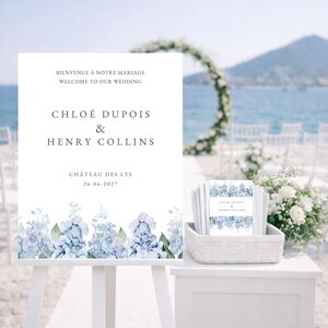 May include: A white wedding sign on an easel, with the text "Bienvenue à Notre Mariage Welcome to Our Wedding" and the names "Chloé Dupois & Henry Collins". The sign features a floral border of blue hydrangeas. A basket of matching cards is visible.