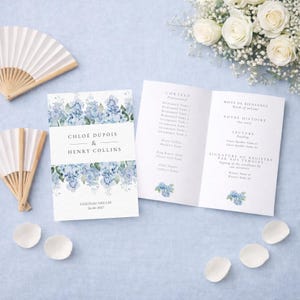 Pode incluir: Conjunto de papelaria de casamento com programa e convite, decorado com motivos florais azuis. O convite diz "Chloé Dupois & Henry Collins" com a data "26-06-2017". Dois leques brancos e pétalas também estão presentes.