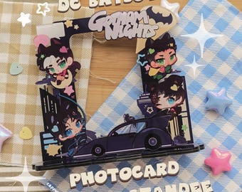 Peut inclure: Un présentoir à photocard en acrylique DC Batboys Gotham Nights avec des personnages de style chibi, une voiture de sport sombre et des silhouettes de ville. Le cadre inclut des étoiles et des cœurs décoratifs, avec le texte DC BATBOYS GOTHAM NIGHTS PHOTOCARD STANDEE imprimé sur la structure.