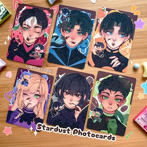 May include: Six Stardust Photocards featuring anime-style illustrations of characters with various hair colors and expressions. Each card has a unique background design, including strawberries, oranges, and blue water droplets. The artist's name, "cappuccineka," is visible on the cards.