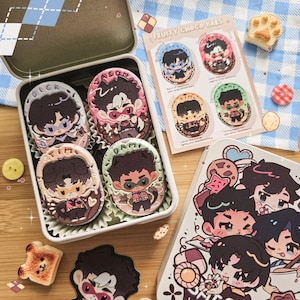 Può includere: Una scatola di metallo piena di sei barrette di cioccolato a tema personaggi. Le barrette sono di forma ovale con glassa colorata e disegni di personaggi. È presente anche una carta con quattro ulteriori disegni di barrette di cioccolato, insieme a una scatola decorativa con illustrazioni di personaggi.