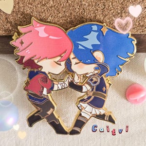 Op de afbeelding: Een goudkleurige emaille pin met twee stripfiguren in een omhelzing. Het ene personage heeft roze haar en de andere blauw haar. De pin heeft het woord "Caitvi" in de rechter benedenhoek.