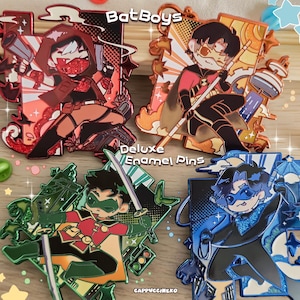 May include: Four rectangular enamel pins featuring stylised cartoon characters in red, orange, green, and blue colour schemes. The pins include text reading BatBoys, Deluxe Enamel Pins, and CAPPUCCINEKO. Each character is depicted in a dynamic pose.