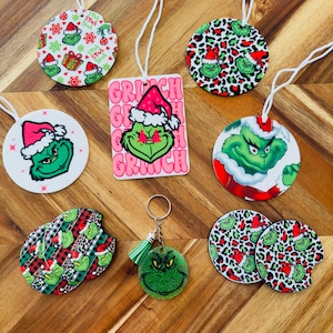 May include: A set of Grinch-themed Christmas ornaments and coasters. The ornaments are round and feature a green Grinch with a red Santa hat. The coasters are round and feature a green Grinch with a red Santa hat and a black and white leopard print. The ornaments and coasters are all made of a white material with a glossy finish. The set also includes a keychain with a green Grinch face and a green tassel.