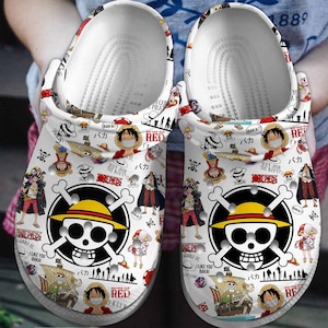Könnte beinhalten: Weiße Clogs mit einem sich wiederholenden Muster aus Anime-Charakteren, Totenköpfen und Schiffen. Das Design umfasst ein großes Totenkopf- und gekreuzte Knochen-Emblem mit einem gelben Hut. Der Text enthält "RED" und japanische Schriftzeichen.