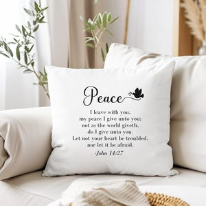 May include: White square throw pillow with the word "Peace" in elegant script, accompanied by a dove illustration. The pillow features a Bible verse from John 14:27 in black text, offering a message of comfort and faith. The pillow is on a white sofa.