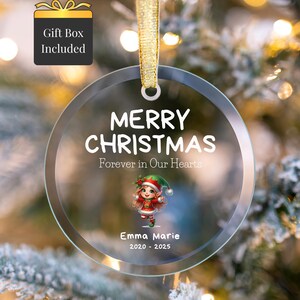 Peut inclure: Une boule de Noël ronde et transparente avec les mots "MERRY CHRISTMAS" et "Forever in Our Hearts" imprimés en blanc. Elle présente un graphisme d'elfe et le nom "Emma Marie" avec les années 2020-2025. La boule est suspendue à un ruban doré.