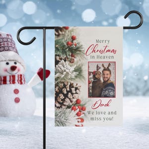 Puede incluir: Una bandera de jardín con temática navideña con un fondo de rama de pino nevada. La bandera presenta una foto de una persona y un perro con cuernos de reno. El texto dice "Feliz Navidad en el cielo, Derek, Te queremos y te echamos de menos!"