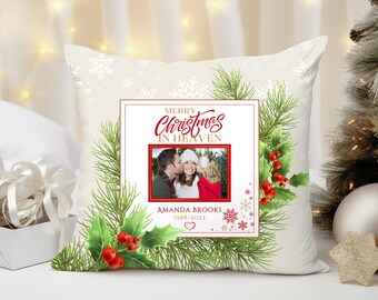 May include: A cream-colored Christmas pillow with a festive design. It features a central framed photo with the text "Merry Christmas in Heaven." The frame is surrounded by holly, pine needles, and red berries. Snowflakes are scattered across the background.