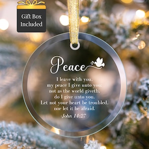 May include: Clear glass Christmas ornament with the word "Peace" in elegant script, accompanied by a dove illustration. The ornament includes a gold ribbon for hanging and features a Bible verse. A gift box is included.