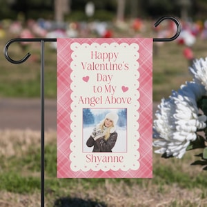 May include: Pink and white garden flag with a plaid pattern and white polka dots. The flag has a photo of a woman in a winter scene and the text "Happy Valentine's Day to My Angel Above" with two pink hearts. The name "Shyanne" is printed at the bottom of the flag.