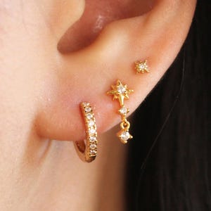 Peut inclure: Ensemble de boucles d'oreilles dorées. Comprend une petite créole, une puce d'oreille en forme d'étoile et un design d'étoile pendant avec des pierres précieuses transparentes. Les boucles d'oreilles sont portées sur l'oreille, mettant en valeur les différents styles.
