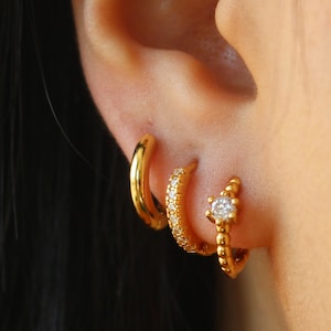 May include: Three gold hoop earrings with small clear stones. The earrings are worn in the earlobe and helix piercing.
