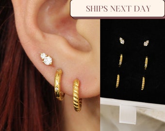 May include: A set of gold hoop earrings with a twisted design and a pair of small stud earrings with clear stones. The earrings are displayed on a black background in a white box with the text "SHIPS NEXT DAY".