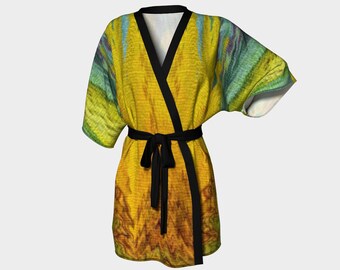 Puede incluir: Una bata estilo kimono amarilla con cinturón y ribete negros. La bata presenta un diseño abstracto colorido con tonos de amarillo, naranja, verde y azul. La bata está hecha de una tela ligera y tiene mangas anchas.