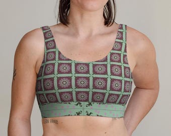 May include: A scoop neck sports bra featuring a repeating square pattern of dark purple and white mandala-like designs on a light green background, with a wide light green band at the bottom decorated with small purple dots and dark silhouettes.