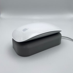 May include: A white wireless mouse with a silver trim sits on a grey charging stand. The mouse has the Apple logo on the top.