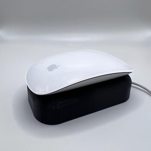 Puede incluir: Un ratón inalámbrico blanco con un logotipo de Apple en la parte superior, descansando sobre una base de carga negra.