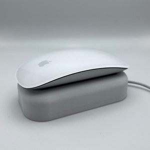 Puede incluir: Un ratón inalámbrico blanco con una base de carga. El ratón tiene un diseño elegante y moderno y presenta el logotipo de Apple en la parte superior.