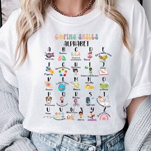 May include: White t-shirt with a colorful alphabet chart featuring coping skills for each letter. The chart includes illustrations and text such as "Ask for help", "Breathing", "Count to 10", "Dancing", "Eat a healthy snack", "Forgive yourself", "Grounding", "Have a friend", "Imagine a peaceful place", "Journal", "Kindness", "Listen to music", "Mindfulness", "Notice the good", "Organize", "Put on music", "Quiet time", "Read a book", "Self-talk", "Take a bath", "Use your senses", "Walk outside", "Write it down", "Yoga", "Zone out and relax".