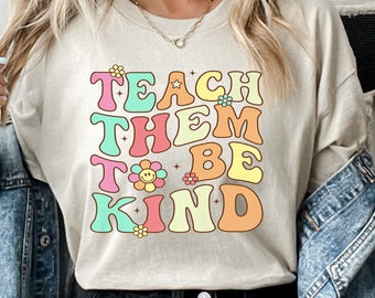 May include: Beige long-sleeve shirt with the text "TEACH THEM TO BE KIND" in colorful, retro-style lettering. The design includes flower and smiley face graphics. The shirt is made of a soft material.