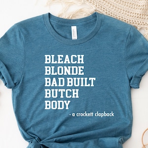 May include: Teal blue t-shirt with white text that reads "Bleach Blonde Bad Built Butch Body - a crockett clapback"
