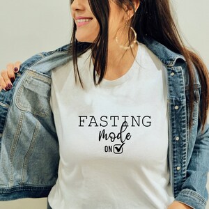 May include: White t-shirt with the text "FASTING mode on" in black script. The shirt is being worn under a denim jacket. The text includes a checkmark symbol.