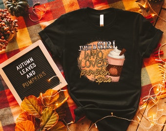 May include: Black t-shirt featuring a graphic of a pumpkin spice latte and the text "Just a girl who loves pumpkin spice". The shirt is lying on a plaid blanket with autumnal decorations.