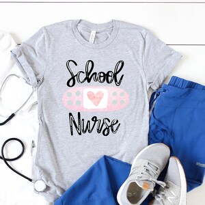 May include: A gray t-shirt with the text "School Nurse" and a pink bandage with a heart on it. The shirt is laid out on a white wooden surface with a stethoscope, a white t-shirt, blue scrubs, and white sneakers.