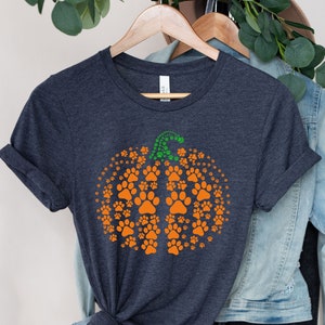 May include: Dark gray t-shirt featuring an orange pumpkin design made of paw prints. The pumpkin has a green stem. The shirt is hanging on a wooden hanger with a denim jacket in the background. A fall-themed graphic tee.