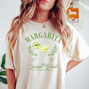 Puede incluir: Camiseta color crema con un dise&ntilde;o de margarita verde. El dise&ntilde;o incluye la palabra "Margarita" sobre una ilustraci&oacute;n de c&oacute;ctel, con "Social Club" y "Save Water Drink Tequila" debajo. La camiseta tiene un corte holgado.