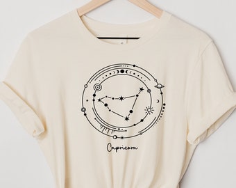 Könnte beinhalten: Ein cremefarbenes T-Shirt mit einer schwarzen Grafik des Sternzeichen Steinbock, umgeben von himmlischen Symbolen. Der Text "Capricorn" ist unter der Grafik gedruckt.