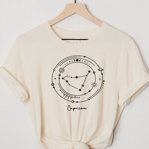 May include: A cream-colored t-shirt with a black graphic of the Capricorn zodiac sign surrounded by celestial symbols. The text "Capricorn" is printed below the graphic.