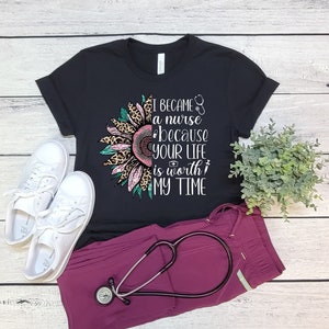 May include: Black t-shirt with a pink and leopard print sunflower graphic. The text "I became a nurse because your life is worth my time" is printed on the shirt. A stethoscope is on a pair of burgundy scrubs.