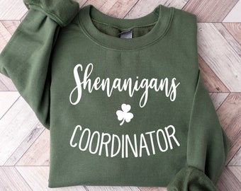 May include: Green sweatshirt featuring the text "Shenanigans Coordinator" in white script, accompanied by a white shamrock. The garment has long sleeves and a crew neck. The background displays a light wood pattern.