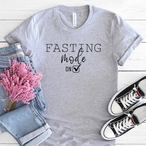 May include: A heather gray t-shirt with the text "FASTING mode ON" in black. The shirt is styled with blue jeans, pink flowers, and black canvas sneakers with white laces, all arranged on a white wooden surface.